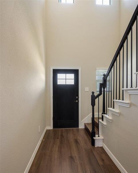 Entryway featuring a black paneled door with glass inserts, wood-finish flooring, a contemporary staircase with black wrought iron spindles, high ceilings, and transom windows Entryway featuring a black paneled door with glass inserts, wood-finish flooring, a contemporary staircase with black wrought iron spindles, high ceilings, and transom windows
