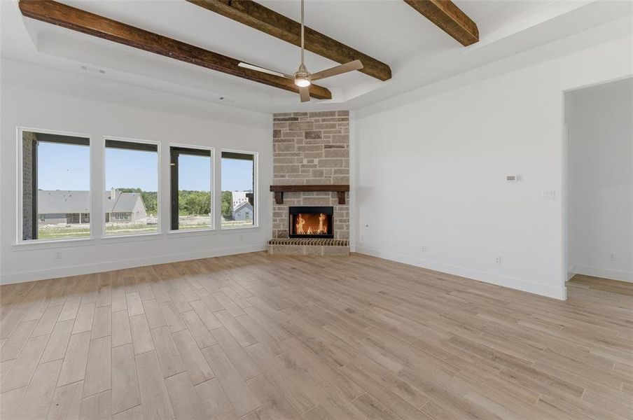 Spacious living area featuring a stone fireplace with a wood mantle, wood-finish flooring, a tray ceiling with exposed wood beams, a ceiling fan, and multiple large windows