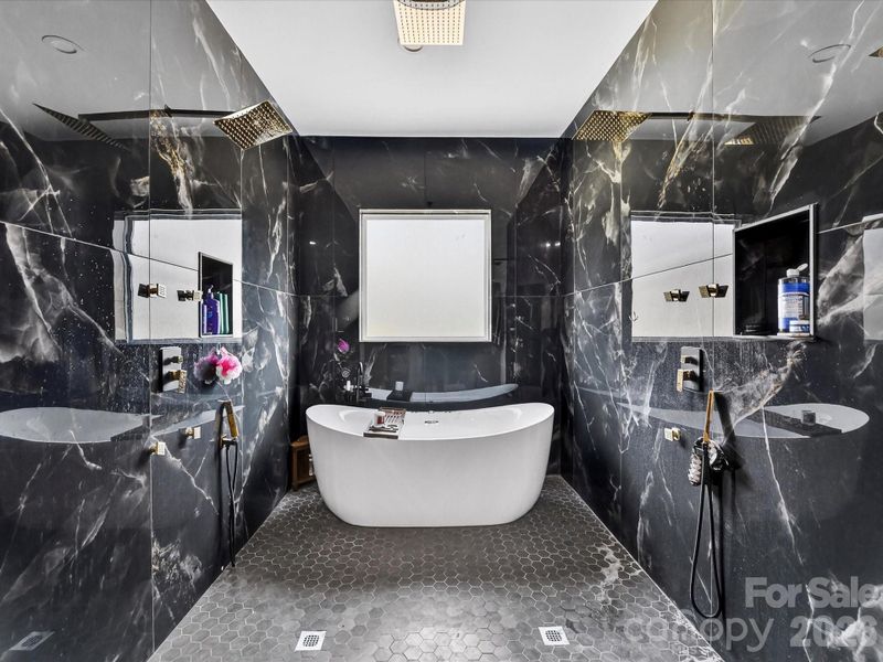 Stunning spa-like bathroom centered around a sleek freestanding soaking tub, surrounded by dramatic marble-style tile and dual rainfall showers.