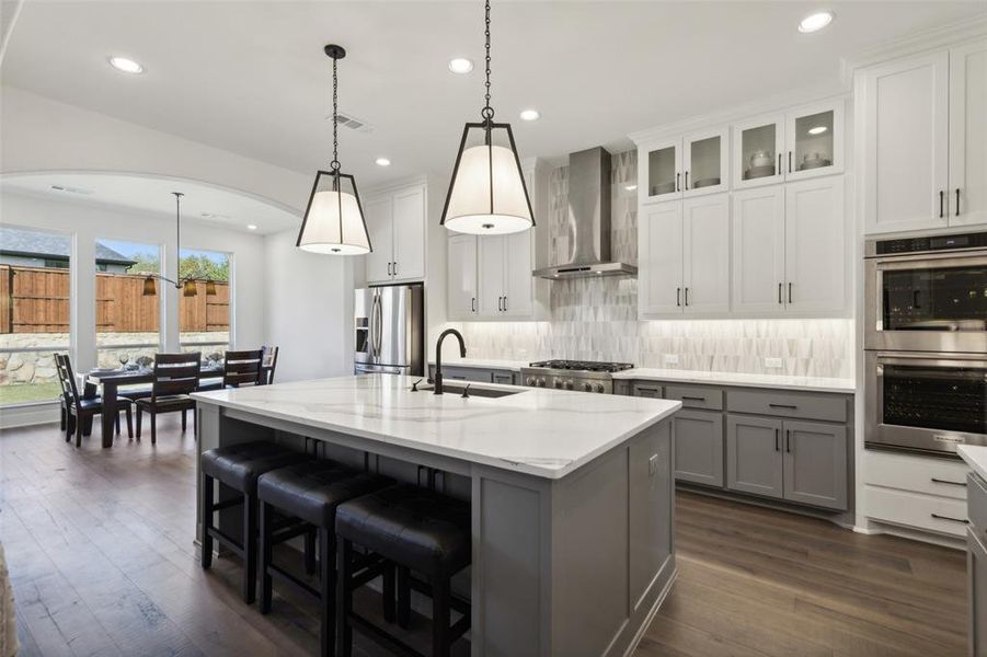 Kitchen featuring wall chimney range hood, an island with sink, hanging light fixtures, white cabinets, and appliances with stainless steel finishes