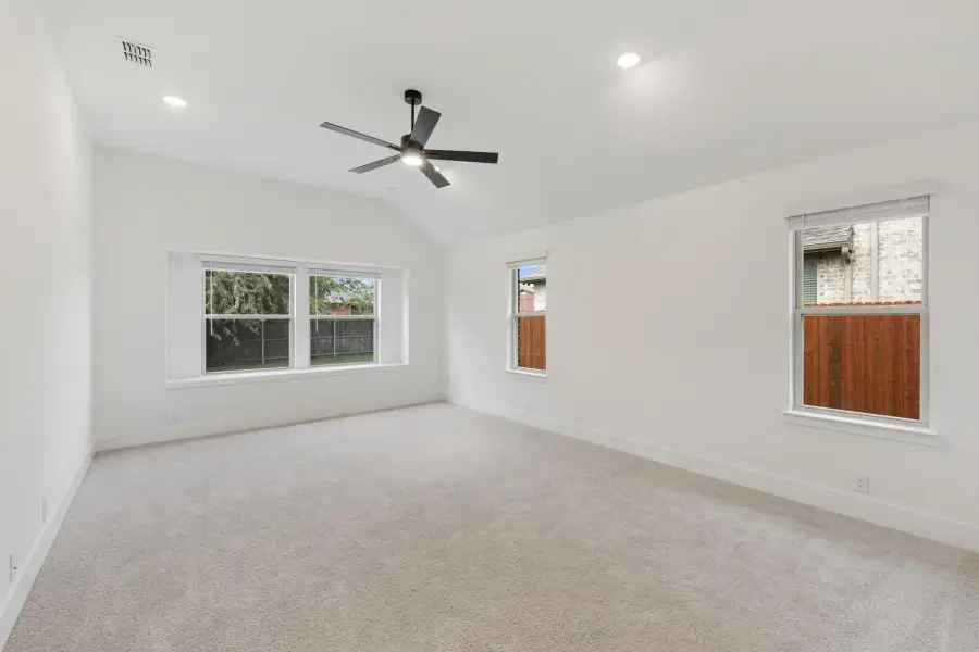 Empty bedroom with vaulted ceiling, black ceiling fan, carpet flooring, and three windows with backyard views