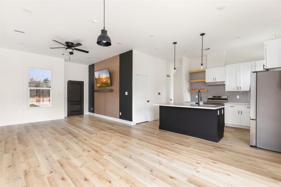 This modern open-concept living space features light wood flooring, a sleek kitchen with white cabinetry, a dark island, and stainless steel appliances. The room is bright, with a large window, stylish fixtures, and a ceiling fan. A mounted TV and accent wall add a contemporary touch.