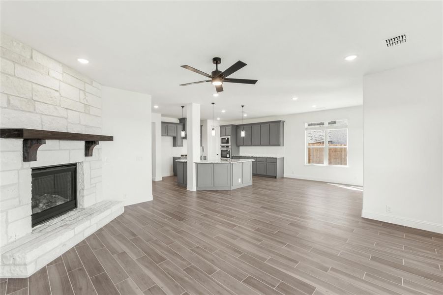 Open living area with stone fireplace, ceiling fan, and open kitchen with gray cabinets and island Open living area with stone fireplace, ceiling fan, and open kitchen with gray cabinets and island