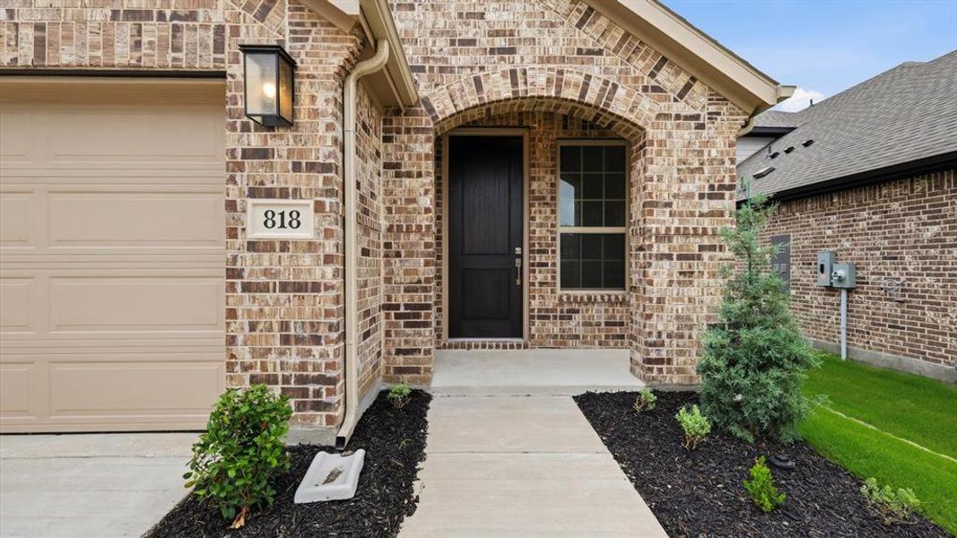 Brick exterior featuring an arched entryway, a dark paneled front door, an exterior wall lantern, a concrete pathway, and a tan garage door