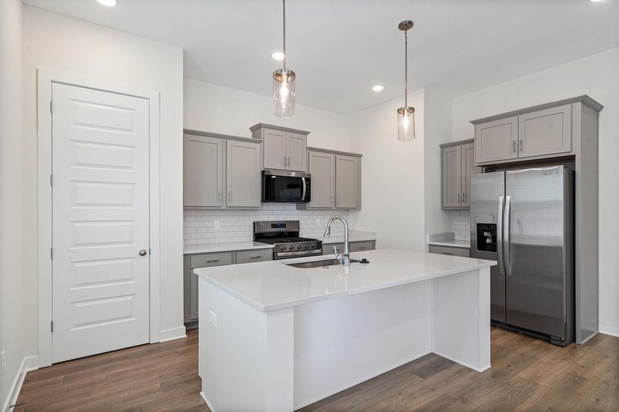 Kitchen featuring gray cabinets, stainless steel appliances, a center island with sink, light stone counters, and decorative light fixtures