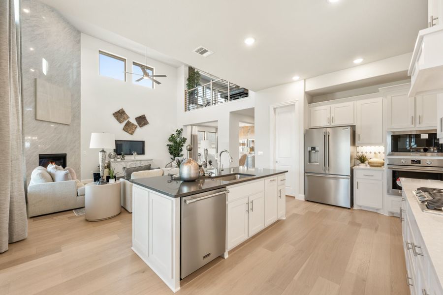 Open-concept kitchen with white cabinets, dark island, stainless steel appliances, and light wood floors opening to living room