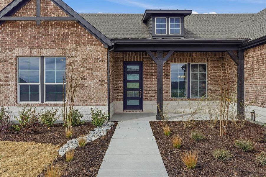 Doorway to property featuring covered porch, a shingled roof, and brick siding