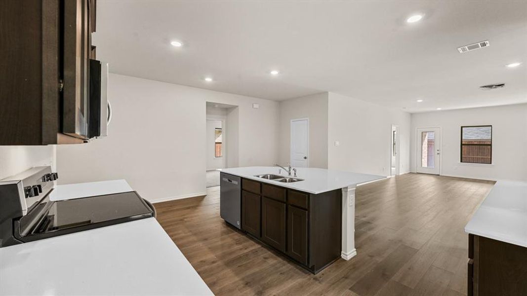 Open-concept kitchen and living area featuring wood-finish flooring, a kitchen island with dual sinks, white countertops, and dark wood cabinetry Open-concept kitchen and living area featuring wood-finish flooring, a kitchen island with dual sinks, white countertops, and dark wood cabinetry