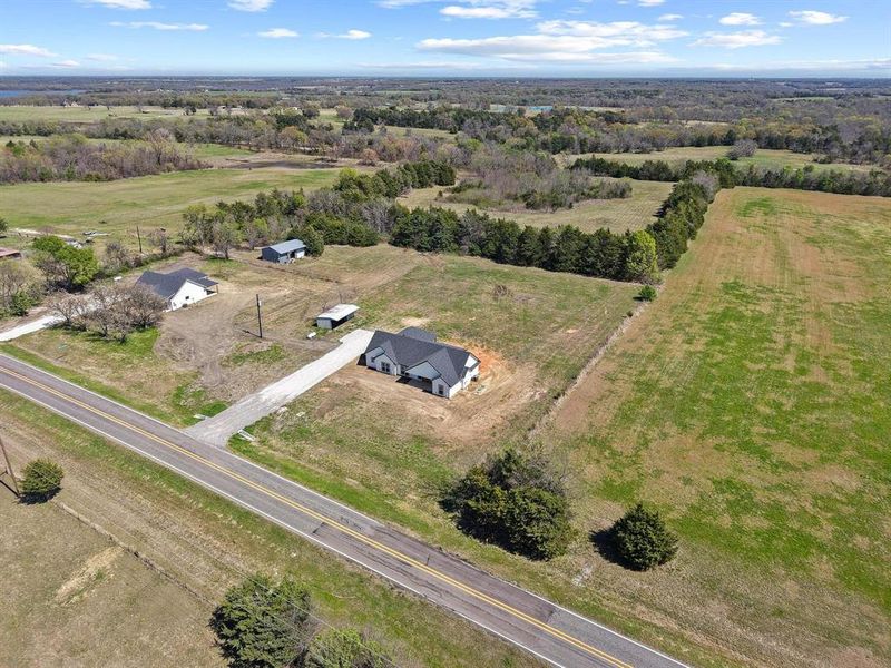 Aerial view showcasing the property's expansive land, including a newly constructed home with a dark roof and a gravel driveway