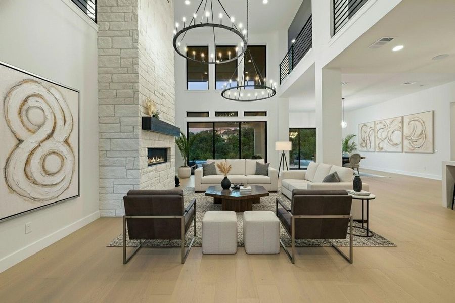 Two-story living area featuring a stone fireplace with a floating mantel, large windows, two circular chandeliers, and wood-finish flooring