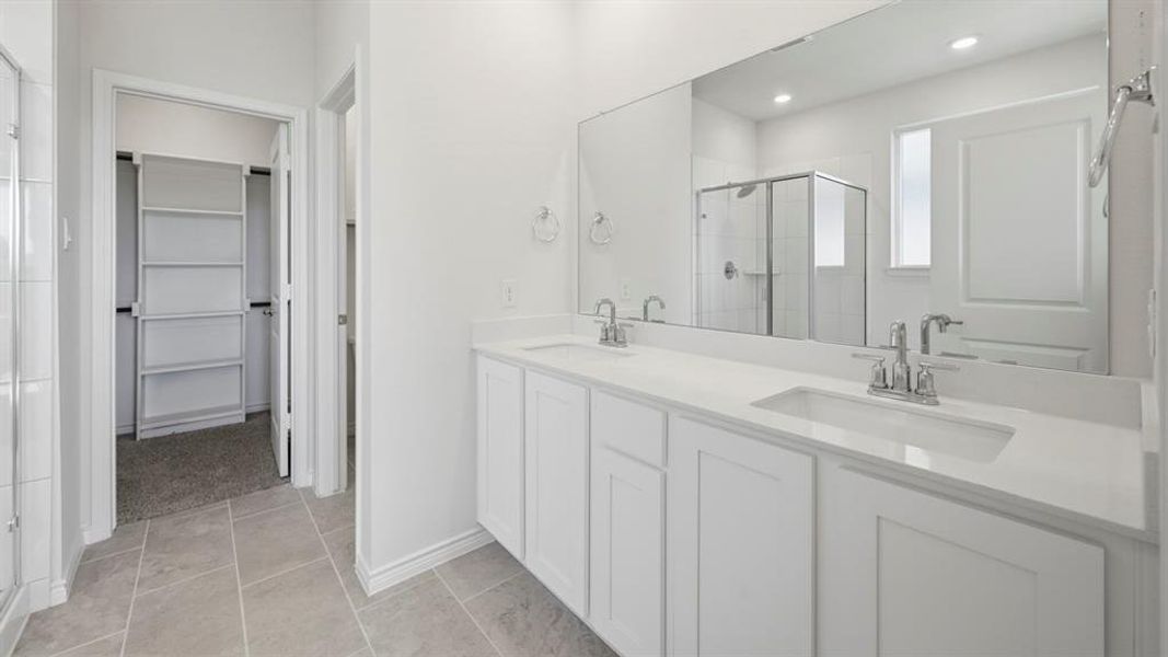 Spacious bathroom featuring a dual vanity with white cabinetry and light-toned countertops, a large frameless mirror, a glass-enclosed shower, and light gray tile flooring Spacious bathroom featuring a dual vanity with white cabinetry and light-toned countertops, a large frameless mirror, a glass-enclosed shower, and light gray tile flooring