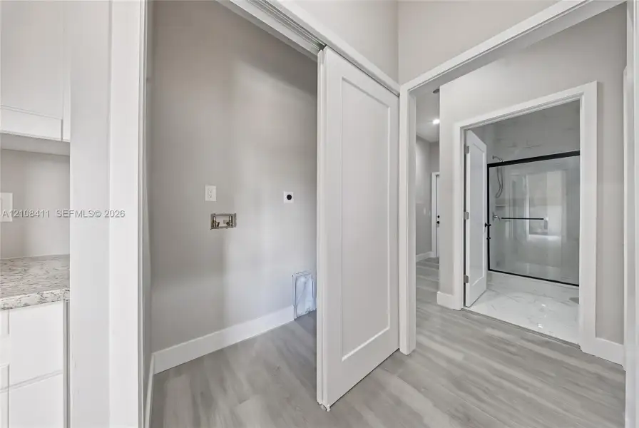 Utility space featuring light-toned wood-finish flooring, white cabinetry with speckled stone-finish countertops, and a sliding door, with a glimpse of a walk-in shower containing light-toned stone-finish tile and a black-framed glass enclosure