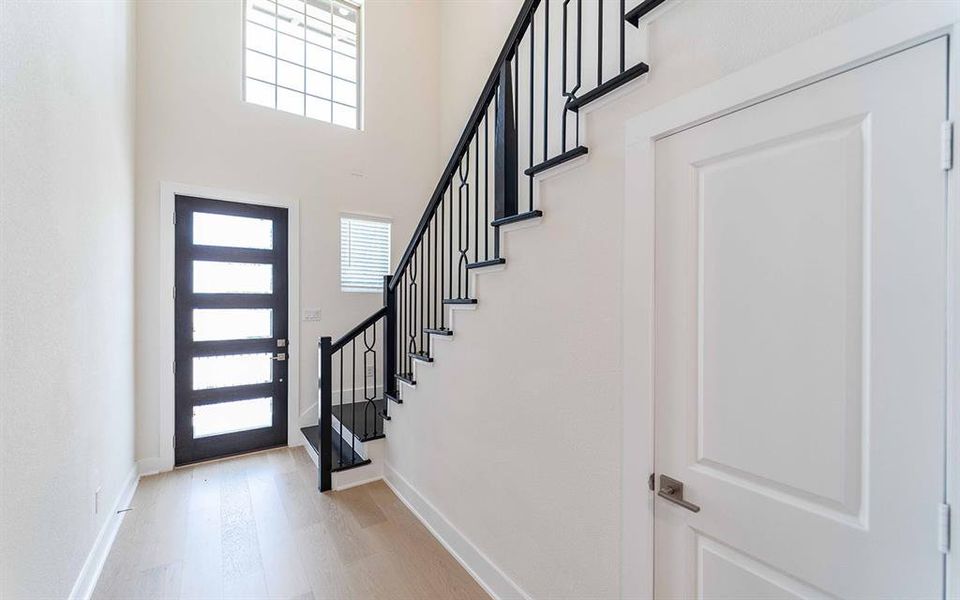 Foyer featuring a modern black entry door with frosted glass panels, light wood-finish flooring, a contemporary staircase with black wrought iron spindles, and a high transom window