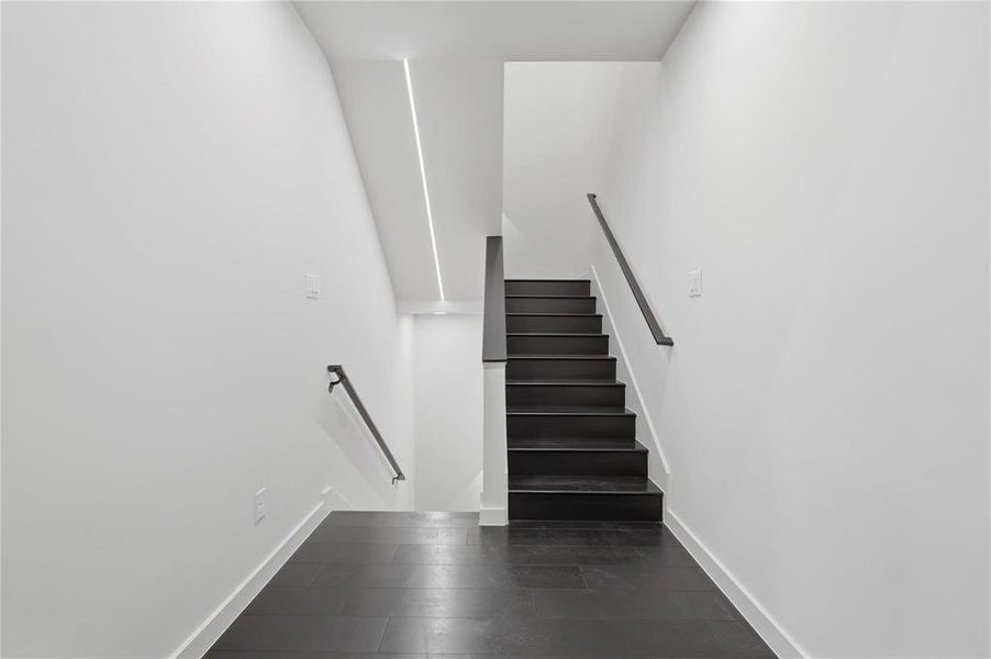 Stairwell featuring dark wood-finish flooring and stairs, white walls, black handrails, and recessed linear ceiling lighting