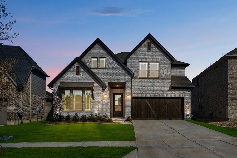 Two-story home with light brick exterior, dark wood garage door, and manicured front lawn at dusk
