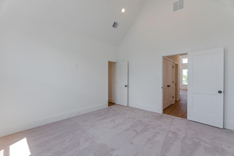 This room features a neutral-colored carpet, high vaulted ceilings, and ample natural light. It includes two doorways, one leading to a hallway and the other to a bright adjoining room with wooden flooring.Photo Disclaimer: Photos are representative of the community and may depict a similar floor plan, model home, or previously completed residence. This room features a neutral-colored carpet, high vaulted ceilings, and ample natural light. It includes two doorways, one leading to a hallway and the other to a bright adjoining room with wooden flooring.Photo Disclaimer: Photos are representative of the community and may depict a similar floor plan, model home, or previously completed residence.