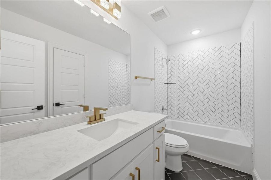 Bathroom featuring a vanity with a light-toned countertop, undermount sink, and brass-finish faucet, accompanied by a tub/shower combination with white subway tile in a herringbone pattern and dark tile flooring