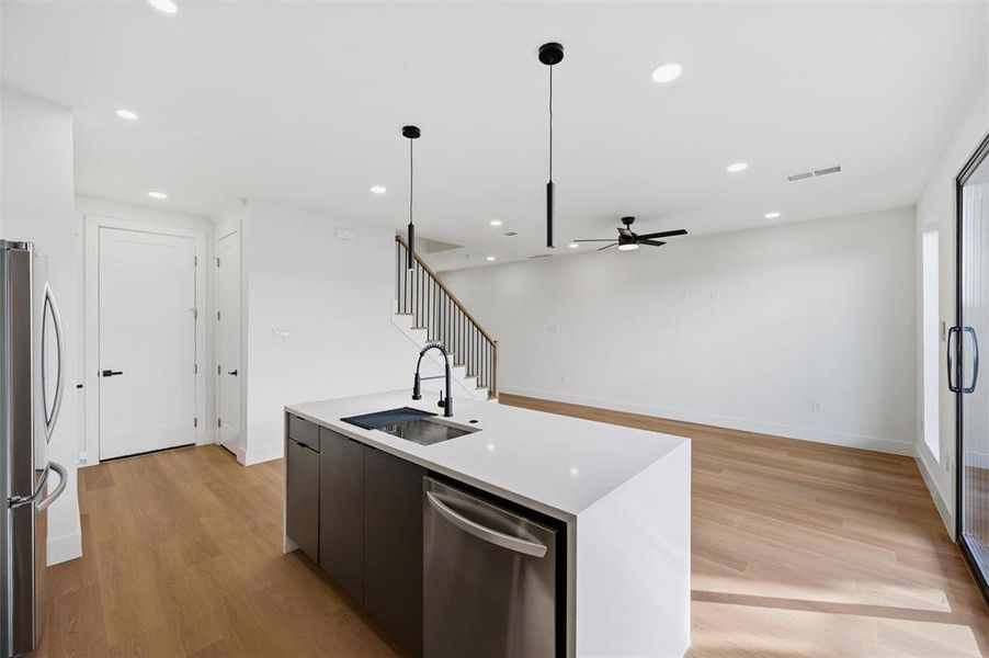 Kitchen features recessed lighting, ceiling fan with light fixture, and pendant lighting above the island. Quartz waterfall island includes a black faucet and built-in dishwasher. Open layout connects directly to the living area and staircase with black metal balusters. Vinyl flooring extends throughout.