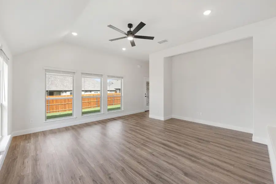Open living room with vaulted ceiling, ceiling fan, wood-look floors, and three backyard-facing windows