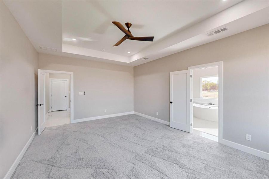 Unfurnished bedroom with connected bathroom, ceiling fan, a raised ceiling, and light colored carpet