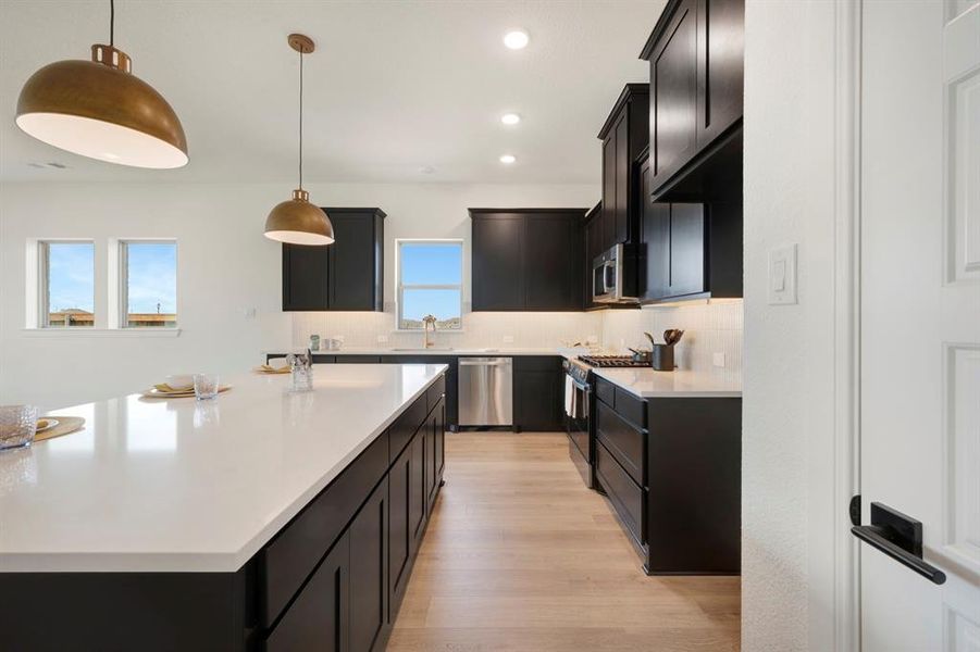 Kitchen featuring dark cabinetry, light wood-style floors, stainless steel appliances, tasteful backsplash, and light stone countertops Kitchen featuring dark cabinetry, light wood-style floors, stainless steel appliances, tasteful backsplash, and light stone countertops