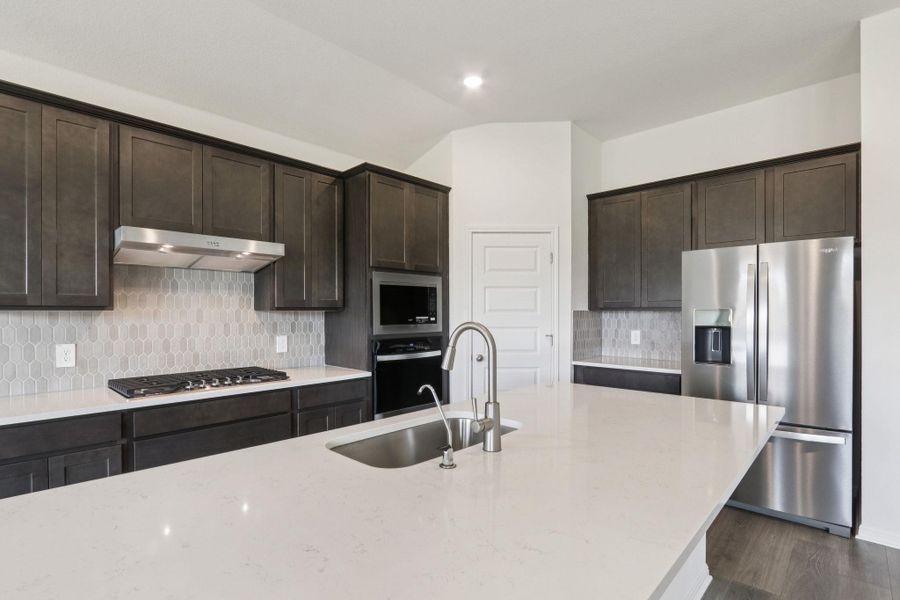 Kitchen featuring dark brown cabinetry, appliances with stainless steel finishes, tasteful backsplash, light stone counters, and under cabinet range hood Kitchen featuring dark brown cabinetry, appliances with stainless steel finishes, tasteful backsplash, light stone counters, and under cabinet range hood