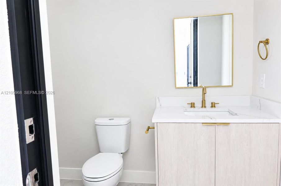 Vanity featuring a light wood-finish cabinet, stone-surface countertop, and gold-toned hardware, accompanied by a rectangular mirror with a gold-toned frame and a gold-toned widespread faucet