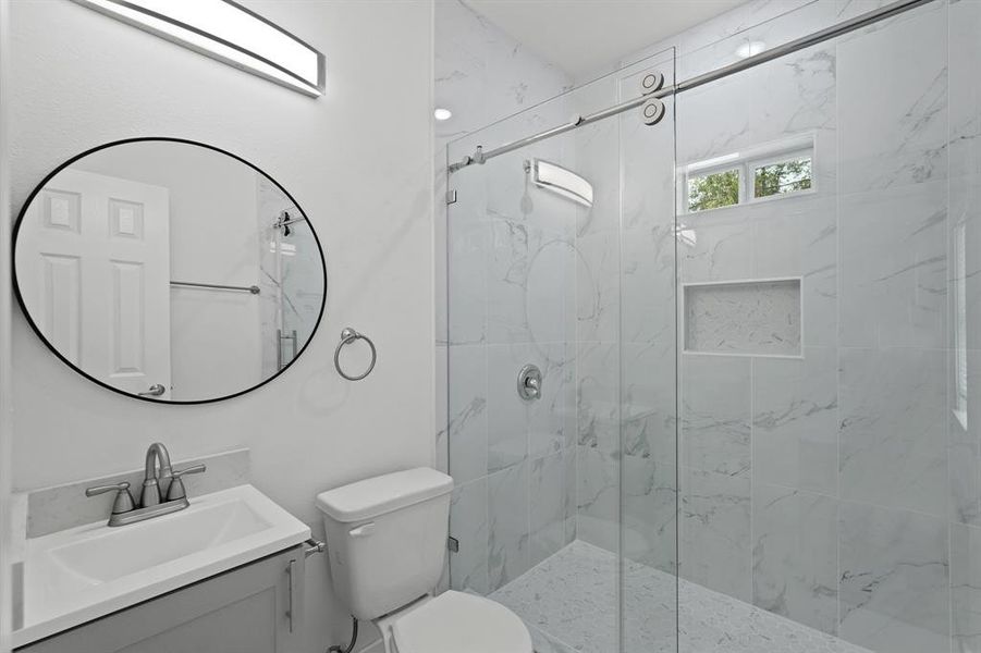 A large walk-in shower with modern tile detailing and recessed lighting, granite countertops, a framed mirror, and wooden cabinetry.