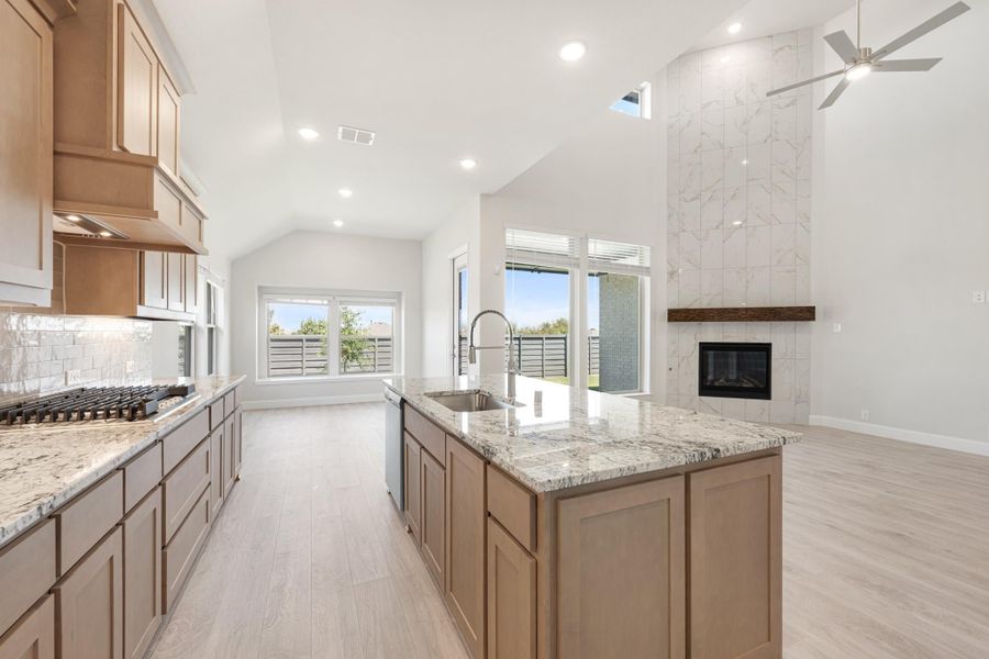 Open kitchen with granite island, wood cabinets, gas cooktop, and vaulted ceiling open to living area with fireplace