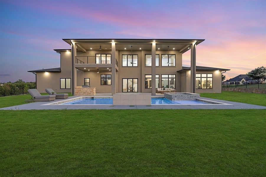 Back of property at dusk with a balcony, a hot tub, stucco siding, a lawn, and an outdoor pool