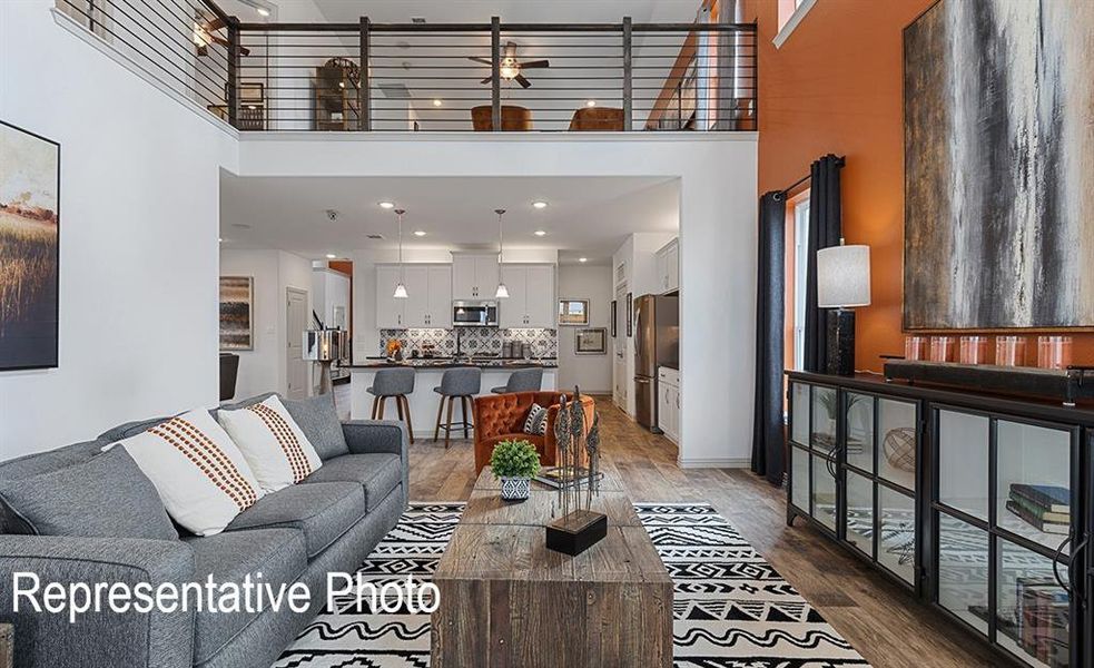 Open-concept living area with high ceilings, wood-finish flooring, a modern kitchen with a breakfast bar, and an upper-level loft with a metal railing