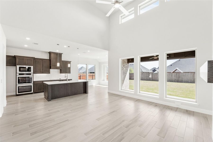 Open-concept kitchen and living area with high ceilings, dark cabinets, center island, and large windows overlooking backyard. Open-concept kitchen and living area with high ceilings, dark cabinets, center island, and large windows overlooking backyard.