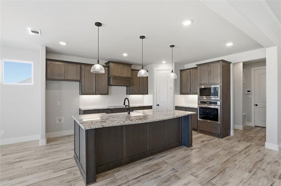 Kitchen with hanging light fixtures, stainless steel appliances, light wood-style floors, light stone countertops, and a center island with sink