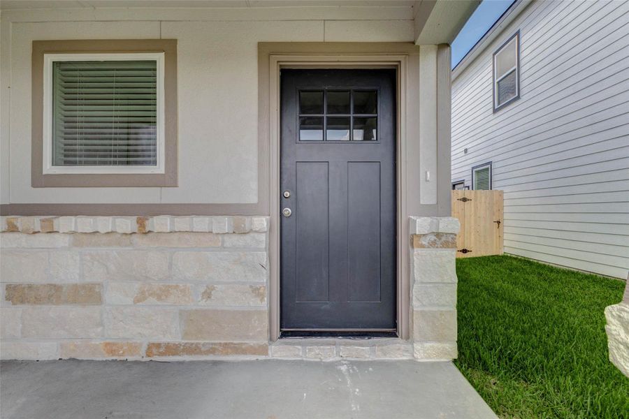 Covered front entry with stone accents, a dark front door, adjacent window, and protected porch area. This close-up highlights the home’s inviting threshold, neutral exterior finishes, and convenient side-yard access with privacy fencing just beyond the entry.