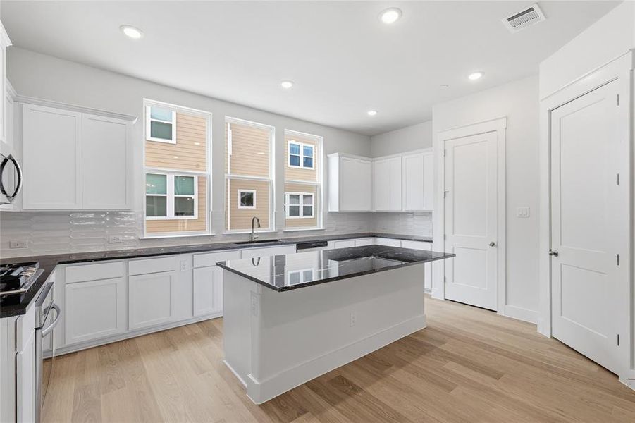 The kitchen features light wood flooring, white cabinetry, a central island with a black countertop, and multiple windows