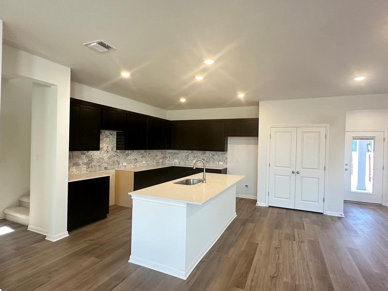Kitchen featuring decorative backsplash, dark wood-style flooring, a kitchen island with sink, light stone countertops, and dark cabinets Kitchen featuring decorative backsplash, dark wood-style flooring, a kitchen island with sink, light stone countertops, and dark cabinets