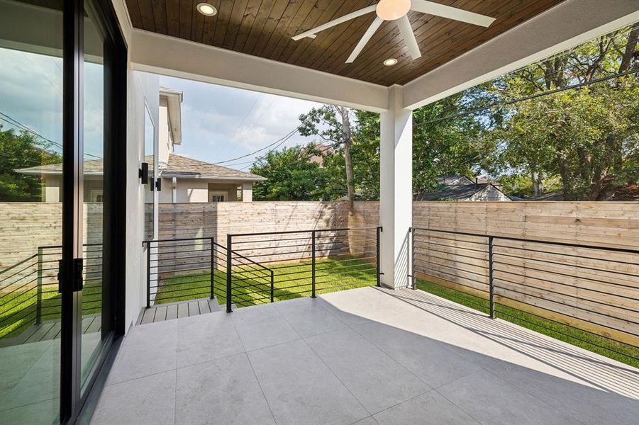 Outside, your custom patio awaits! Features include tile floors, a metal railing that offers a sleek and minimalistic aesthetic, and a large ceiling fan that hangs from the center accenting the wood planks overhead.