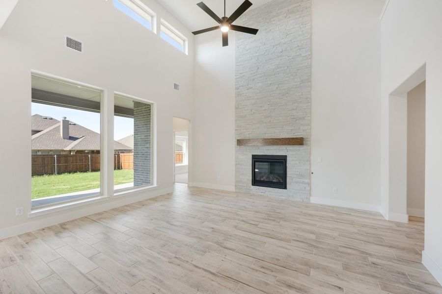Open living room with high ceilings, stone fireplace, ceiling fan, and large windows overlooking backyard Open living room with high ceilings, stone fireplace, ceiling fan, and large windows overlooking backyard