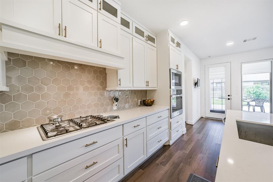 A gas cooktop paired with a vented hood, complemented by a sleek tile backsplash, and featuring a built-in stainless steel oven and microwave for a seamless, modern kitchen design. A gas cooktop paired with a vented hood, complemented by a sleek tile backsplash, and featuring a built-in stainless steel oven and microwave for a seamless, modern kitchen design.