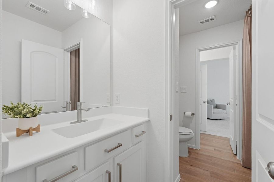 This photo showcases a bright, modern bathroom with a large mirror, white vanity, and sleek fixtures. It features wood-style flooring, a visible toilet, and connects to a well-lit room beyond.