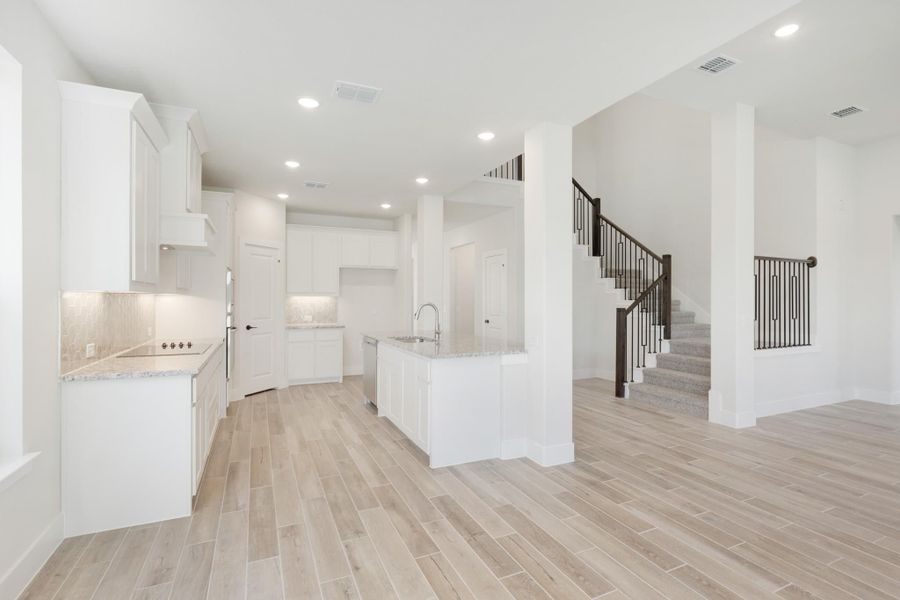 Open kitchen with white cabinets, granite island, and wood-look tile floors, open to staircase with iron railing