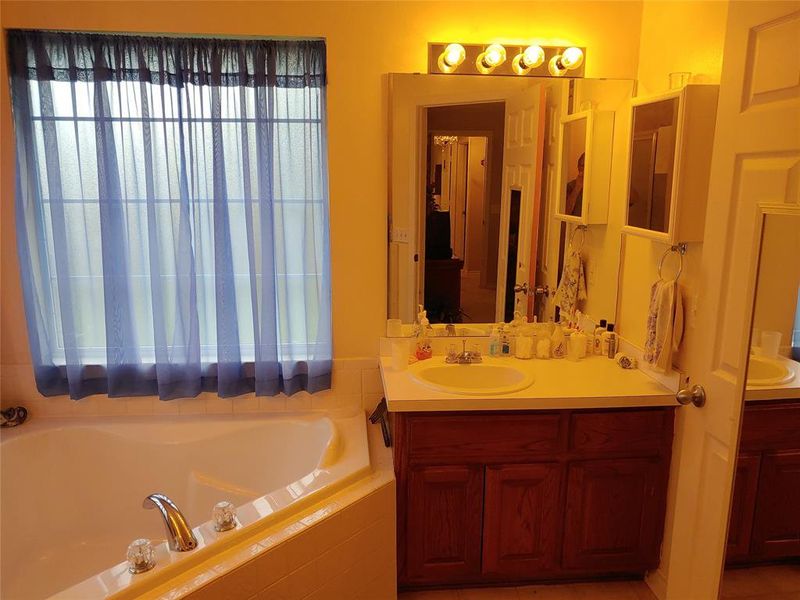 Bathroom featuring a corner soaking tub with chrome fixtures, a window with sheer blue curtains, and a vanity with a single basin sink and wood-finish cabinetry
