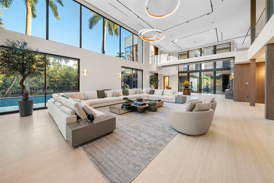 Expansive double-height living room with oversized curved sectional, ring chandelier, floor-to-ceiling glass with direct pool and tropical garden views, open second-floor walkway with glass railing, and seamless flow to foyer and kitchen.