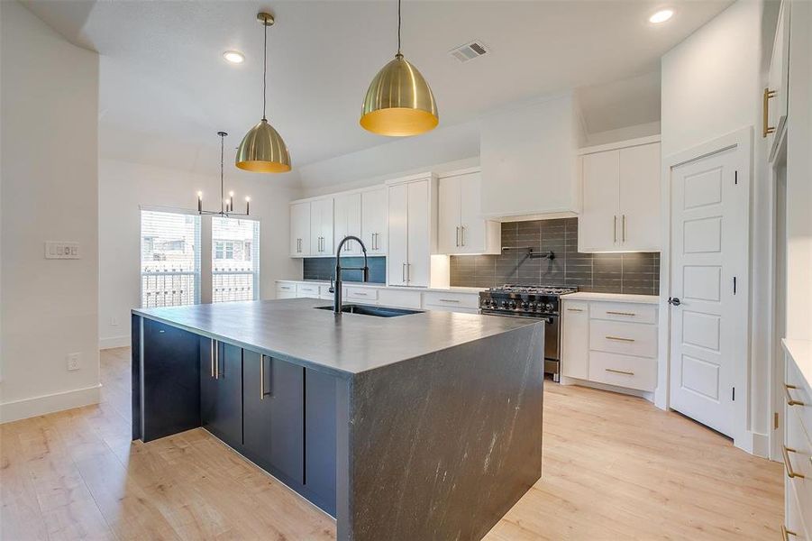 Kitchen featuring light hardwood / wood-style floors, hanging light fixtures, a kitchen island with sink, stainless steel range, and tasteful backsplash