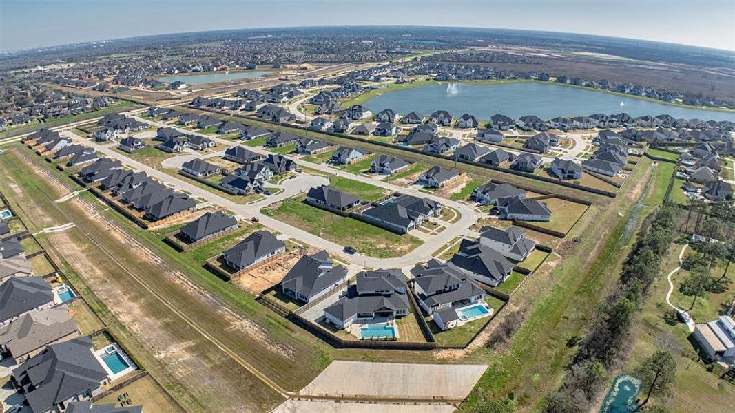 This aerial photo showcases a suburban neighborhood with modern homes, many featuring private pools. The community is organized in a grid-like pattern, with wide streets and ample green spaces. A nearby lake adds a scenic element, offering potential recreational opportunities and enhancing the overall attractiveness of the area.