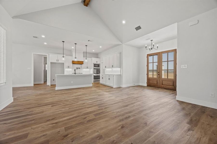 Unfurnished living room featuring beam ceiling, light wood-style flooring, a chandelier, french doors, and high vaulted ceiling Unfurnished living room featuring beam ceiling, light wood-style flooring, a chandelier, french doors, and high vaulted ceiling