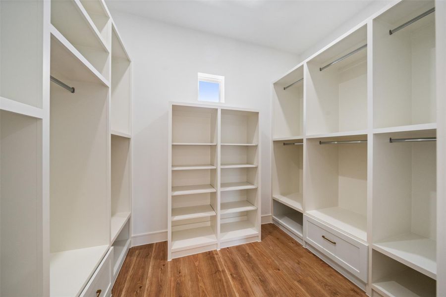 Spacious boutique style walk-in closet features ample shelving and hanging space, bright white cabinetry, and a stylish wooden floor, offering a clean and organized look.