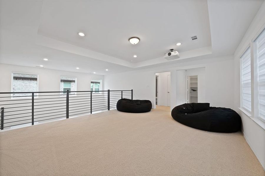 Carpeted loft area featuring a tray ceiling with recessed lighting, built-in projector, white window blinds, and contemporary horizontal bar railing