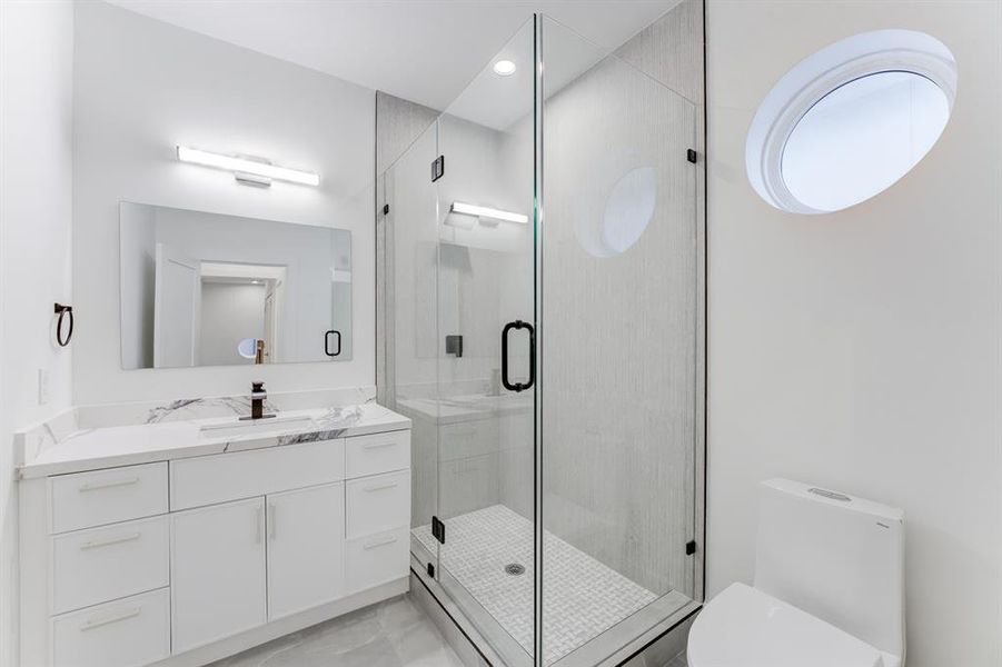 Circular window, frameless shower door and mirror, quartz countertops, and chic white cabinetry for storage.