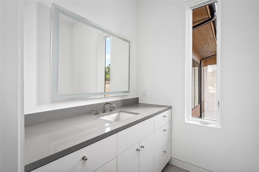 This stylish secondary ensuite bath features sleek gray quartz countertops that seamlessly extend into the matching quartz shower surround, creating a cohesive and modern design. A pocket door maximizes space, while the front-lit mirror adds a touch of elegance and functionality.